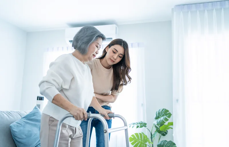 Adult daughter steadying her mother as she walks with a mobility walker at home
