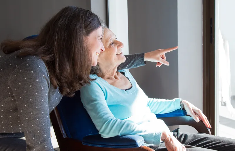 Adult daughter and her senior mother looking out a window together in a residential senior care home