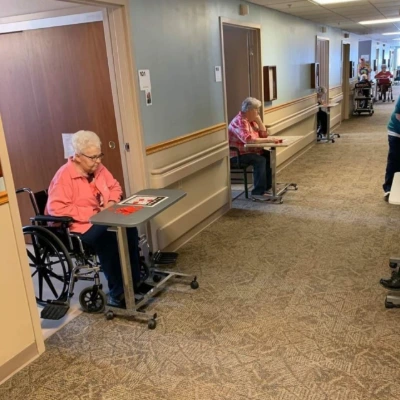 Residents of a care community seated at individual tables spaced along a bright hallway