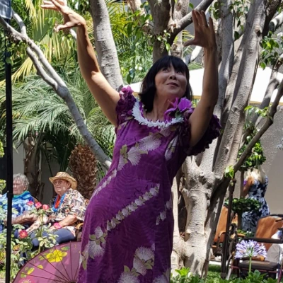 Senior woman in a purple dress and flower lei dancing hula outdoors as other residents watch