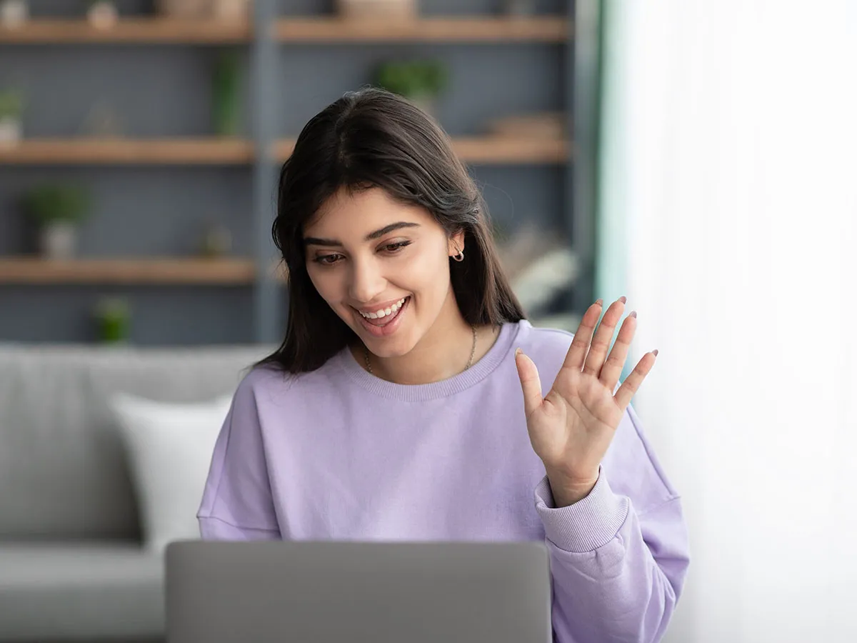 Woman in a lavender sweater waving warmly at her laptop during a video call