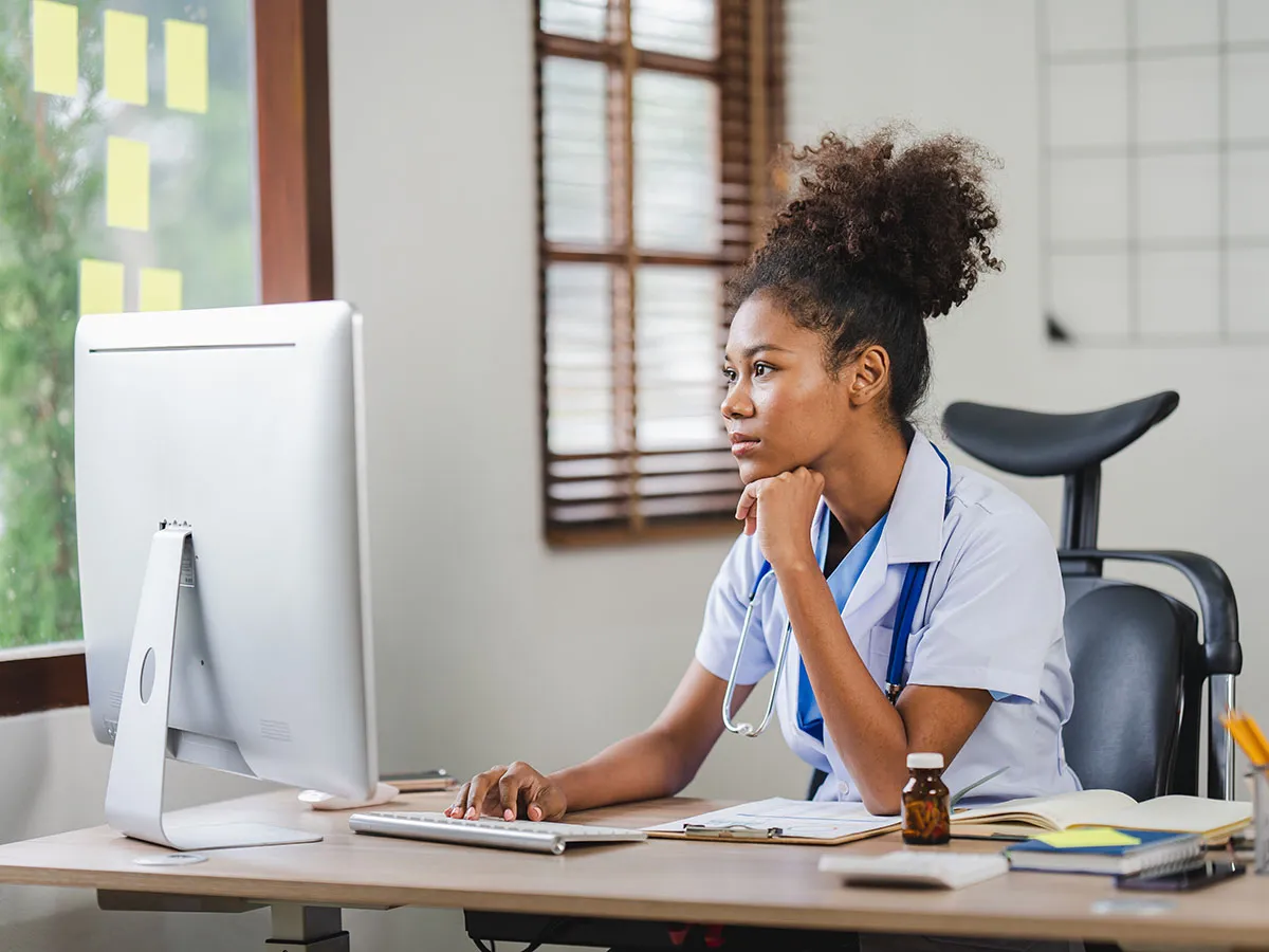 Physician at her desktop computer with a stethoscope, reviewing a patient chart in a bright office