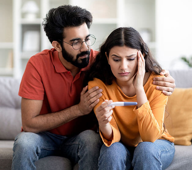 A sad couple on a couch. The man comforts the woman; she's holding a pregnancy test and looks worried.

